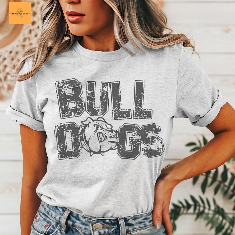 Puede incluir: Una camiseta gris claro con la palabra "BULLDOGS" en letras grises desgastadas. La camiseta presenta un gr&aacute;fico de bulldog. La camiseta la lleva una persona.