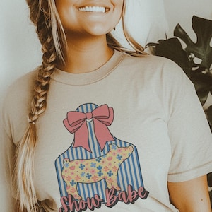 May include: Beige t-shirt featuring a graphic of a pig with a floral pattern, a pink bow, and blue stripes. The text "Show Babe" is written in a script font below the pig. The shirt is a crew neck.