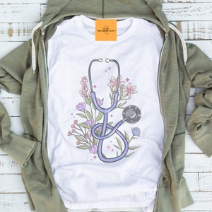 May include: White t-shirt with a blue stethoscope and pink, purple, and blue flowers. The text "marigold lane" is on a yellow tag.