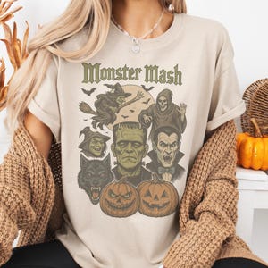 May include: Beige t-shirt with a Halloween-themed graphic. The design features the text "Monster Mash" above illustrations of a witch, a grim reaper, Frankenstein's monster, a werewolf, and jack-o'-lanterns. The artwork is in shades of green, orange, and black.