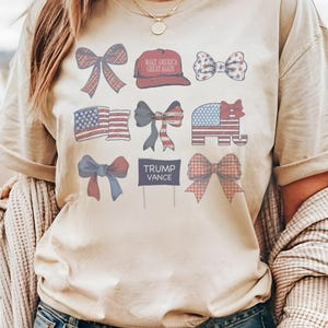 May include: A beige t-shirt with a red, white, and blue design. The design includes a red baseball cap with the text "Make America Great Again", a red, white, and blue bow, an American flag, a red, white, and blue elephant, a red, white, and blue bow, a sign with the text "Trump Vance", and a red, white, and blue bow.