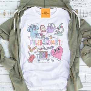 May include: White t-shirt with a colorful graphic design featuring medical-themed illustrations, including a stethoscope, syringe, and heart. The text "Phlebotomist love" is prominently displayed. The shirt is partially visible, with a green zip-up hoodie surrounding it.
