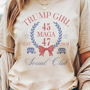 May include: A beige t-shirt with a blue and red graphic design that says "Trump Girl 45 MAGA 47 2024 EST" with a red bow and two blue elephant silhouettes with stars. The text "Social Club" is written in cursive below the graphic.