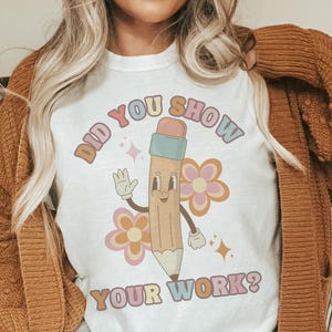 May include: White t-shirt with the text "DID YOU SHOW YOUR WORK?" in retro-style lettering. The shirt features a cartoon pencil character waving, with flowers and sparkles. The pencil has a blue eraser and a light brown body.