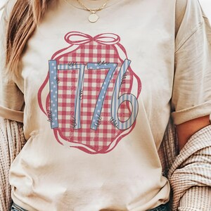 May include: Beige t-shirt featuring a graphic design with the number "1776" in blue and red. The "1776" design is set against a red and white gingham pattern, with a red ribbon-like border.