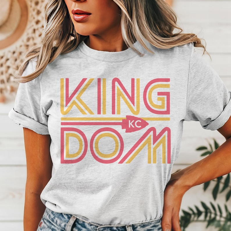 May include: A light gray t-shirt with the word "KINGDOM" in yellow and pink block letters. The word "KC" is in the center of the shirt, with an arrow pointing to the right. The shirt is made of a soft material.