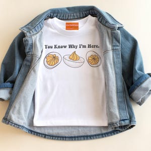 May include: A white t-shirt with the text "You Know Why I'm Here." and a graphic of deviled eggs. The shirt is displayed inside a light blue denim jacket.