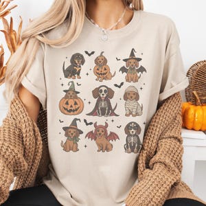 May include: Beige t-shirt featuring a Halloween-themed design with dachshunds dressed in various costumes. The dogs are depicted as a pumpkin, a witch, a mummy, a skeleton, and a bat. Black bats are scattered around the dogs.