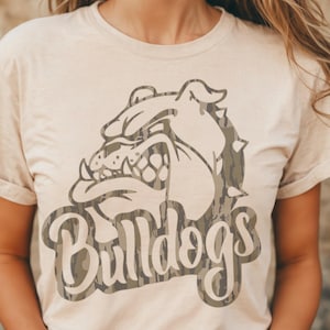 May include: A beige t-shirt featuring a bulldog graphic with a camouflage pattern. The word "Bulldogs" is printed in a stylized font below the bulldog image. The shirt has short sleeves and a crew neck.