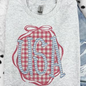 May include: A light gray t-shirt featuring a red and white gingham design with the word "USA" in blue and white stripes. The graphic is framed by a red outline and bow.