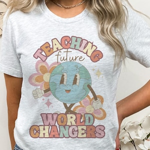 May include: Light gray t-shirt with the text "TEACHING future WORLD CHANGERS" in a retro font. The graphic features a smiling globe with arms and legs, surrounded by flowers and stars.