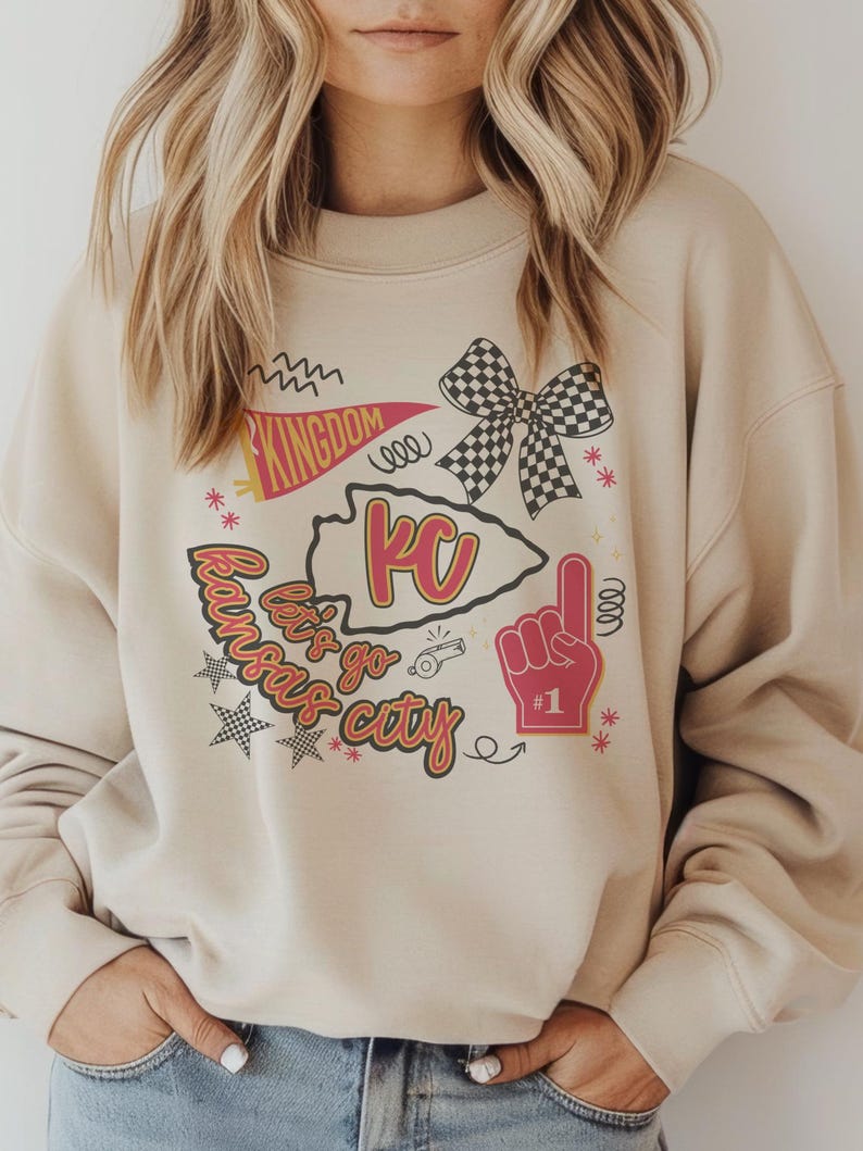 May include: A beige sweatshirt with a graphic design featuring the text "Kansas City" and "Kingdom". The design includes a KC arrowhead, a checkered bow, a foam finger, and stars. The sweatshirt is a neutral color.