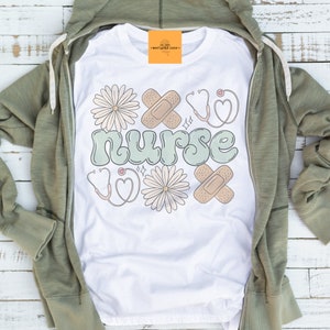 May include: White t-shirt with a green "nurse" graphic. The graphic is surrounded by white daisies, band-aids, and red stethoscopes.