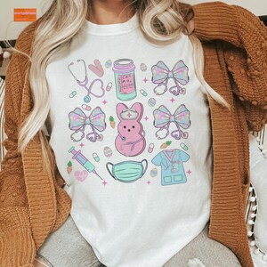 May include: White t-shirt with a pastel pink and blue Easter-themed design. The design features a pink Peeps bunny with a nurse's hat and stethoscope, a syringe, a face mask, a bottle of pills, bows, and Easter eggs.
