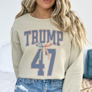 May include: A beige sweatshirt with the text "TRUMP 47" printed in blue and red. A red, white, and blue ribbon is above the number 47.