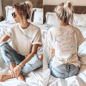 May include: A white t-shirt with a red, white, and blue ribbon design and the text "Land of the free, because of the brave" printed on the back. The shirt is tied at the back.