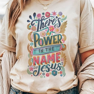 May include: A beige t-shirt with a colorful graphic design that says "There's Power in the Name of Jesus".