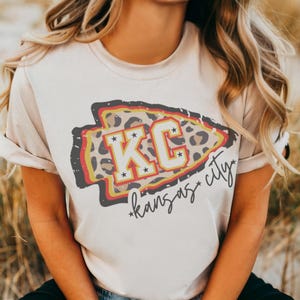May include: A white t-shirt with a graphic of a red, yellow, and black arrow with the letters "KC" in white and the text "*kansas city*" in black script.