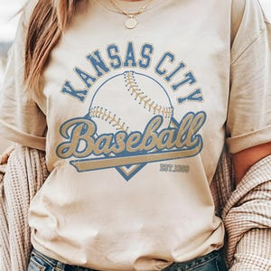 May include: Cream-colored t-shirt featuring a baseball design with the words "Kansas City Baseball" in blue and gold lettering. The shirt includes the text "EST. 1969" below the baseball graphic. The shirt has short sleeves.