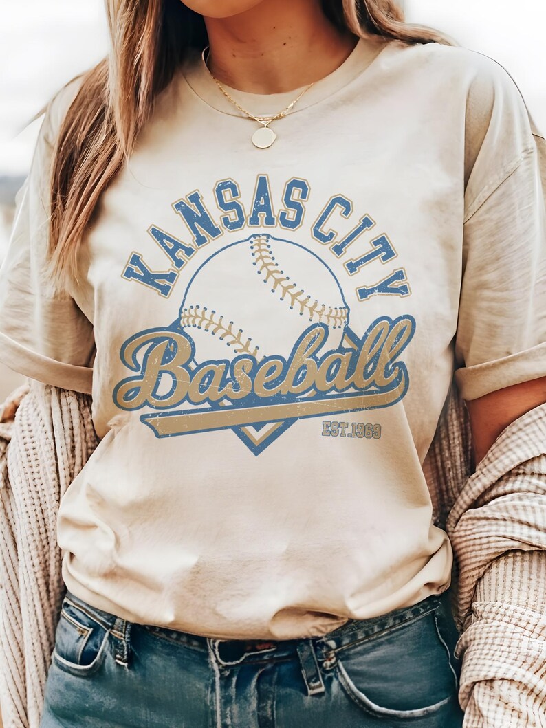 May include: Cream-colored t-shirt featuring a baseball design with the words "Kansas City Baseball" in blue and gold lettering. The shirt includes the text "EST. 1969" below the baseball graphic. The shirt has short sleeves.
