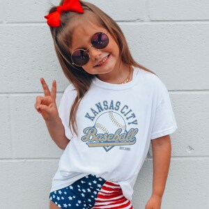 May include: A young person wearing a white t-shirt with "Kansas City Baseball" text and a baseball graphic. They are also wearing blue shorts with white stars and red and white striped shorts. A red bow is in their hair.