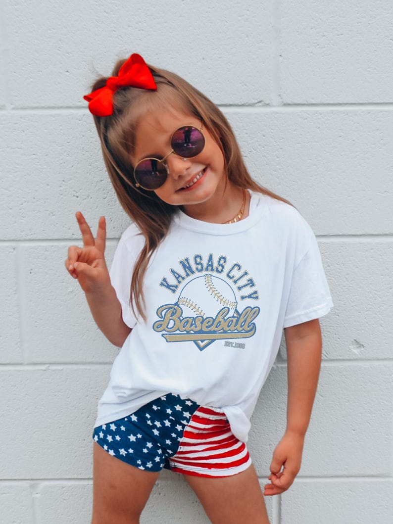 May include: A young person wearing a white t-shirt with "Kansas City Baseball" text and a baseball graphic. They are also wearing blue shorts with white stars and red and white striped shorts. A red bow is in their hair.