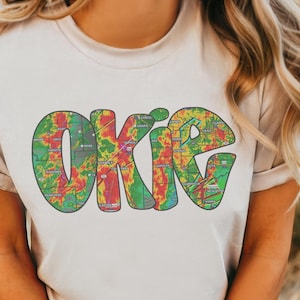 May include: Cream-colored t-shirt featuring the word "OKIE" in large, colorful letters. The letters are filled with a map-like design in shades of green, red, yellow, and blue. The shirt has short sleeves and a relaxed fit.