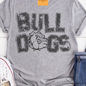 Puede incluir: Una camiseta gris jaspeado con la palabra "BULLDOGS" en letras negras desgastadas y un gr&aacute;fico de bulldog. La camiseta est&aacute; doblada con unos vaqueros azules y una zapatilla blanca.