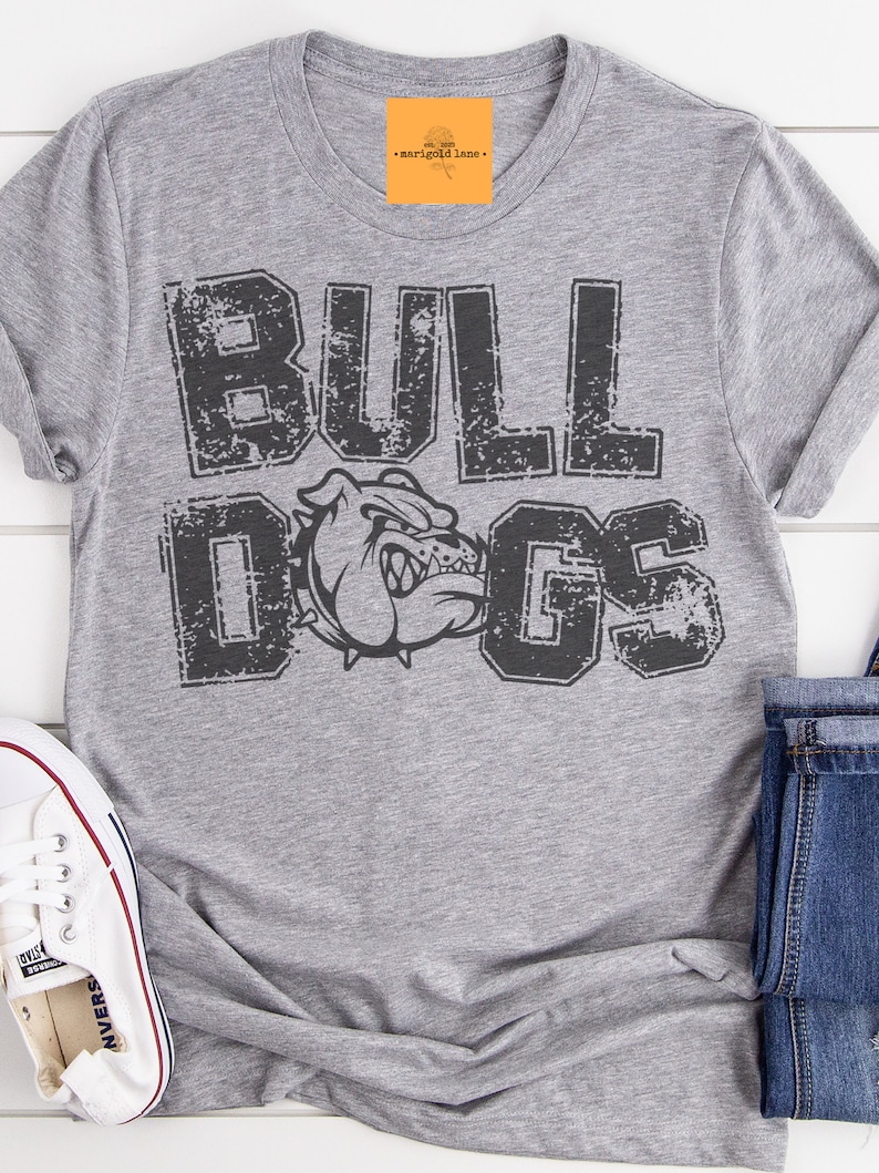 Puede incluir: Una camiseta gris jaspeado con la palabra "BULLDOGS" en letras negras desgastadas y un gr&aacute;fico de bulldog. La camiseta est&aacute; doblada con unos vaqueros azules y una zapatilla blanca.