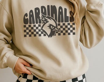Puede incluir: Sudadera beige con la palabra "CARDINALS" en letras oscuras en bloque sobre un patrón de tablero de ajedrez y un gráfico de pájaro cardenal. La sudadera se combina con pantalones a cuadros blancos y negros.