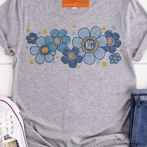 May include: Gray t-shirt with a blue and gold floral design. The flowers have a retro style and the center flower has the letters "KC" in the middle.