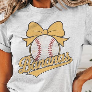 May include: A white t-shirt with a yellow baseball with a red stitching and a yellow bow on top. The text "Bananas" is written in yellow with a yellow outline.