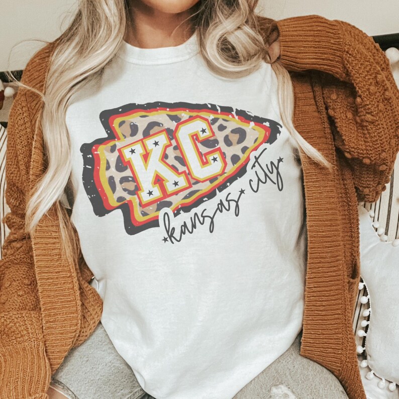 May include: White t-shirt with a graphic of a leopard print arrow with the text "KC*kansas city*" in black.