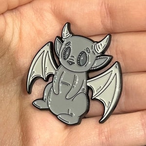 Könnte beinhalten: Eine graue Emaille-Anstecknadel mit einer Karikatur einer geflügelten Kreatur mit großen Augen und Hörnern. Die Kreatur sitzt aufrecht mit ausgebreiteten Flügeln hinter sich.