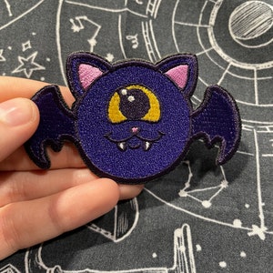May include: Purple and pink embroidered bat with a single large yellow eye and small fangs.