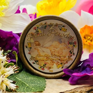 May include: A round, brown container with a gold lid and label. The label reads "Petal Powder Rose Petal & Mineral Oil Shimmering Highlight" and features a floral design with a fairy.