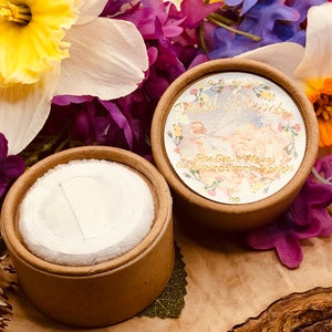 May include: Two round cardboard containers with lids. The container on the left is empty and the container on the right has a label that says "Bardot Beauty - Petal Powder - Rose Glow & Magical Shine - Soft & Smooth Finish". The containers are surrounded by purple, yellow, and white flowers.