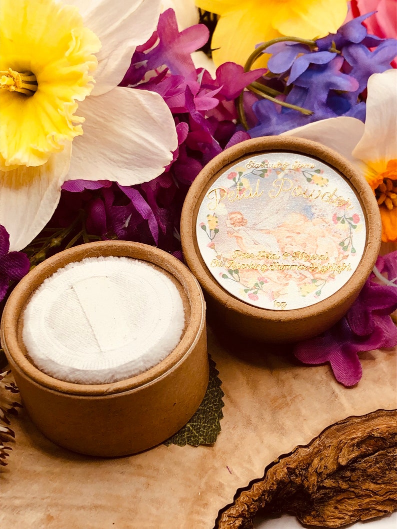 May include: Two round cardboard containers with lids. The container on the left is empty and the container on the right has a label that says "Bardot Beauty - Petal Powder - Rose Glow & Magical Shine - Soft & Smooth Finish". The containers are surrounded by purple, yellow, and white flowers.