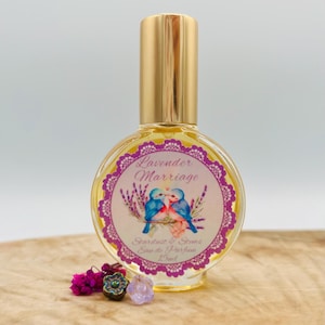 May include: A round perfume bottle with a gold cap, filled with a yellow liquid. The label reads "Lavender Marriage" with a design of two blue birds. The bottle is 15ml and is from "Stardust & Scents".