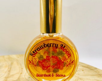 May include: A small glass bottle with a gold cap and a round label that says "Strawberry Moon Stardust & Stems 15ml". The label has a red and yellow illustration of strawberries.