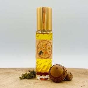 May include: A clear glass rollerball bottle filled with yellow liquid, featuring a gold cap and a tan label with the text Cottage Witch, Stardust & Stone, 10ml. The bottle is decorated with a small cluster of green moss and two brown acorns.