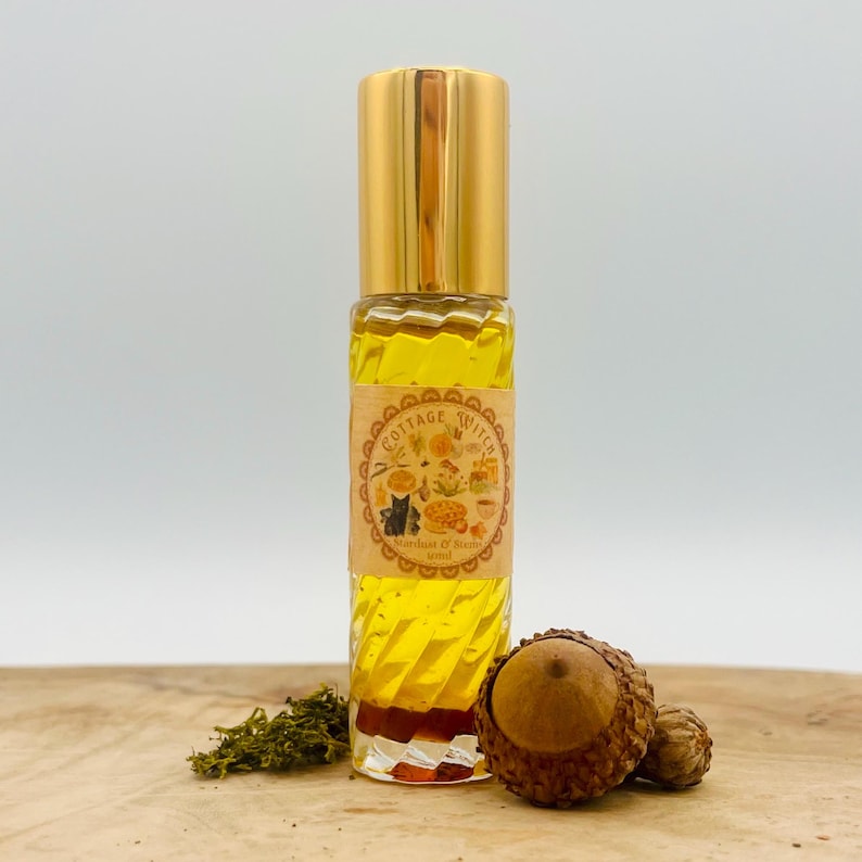 May include: A clear glass rollerball bottle filled with yellow liquid, featuring a gold cap and a tan label with the text Cottage Witch, Stardust & Stone, 10ml. The bottle is decorated with a small cluster of green moss and two brown acorns.
