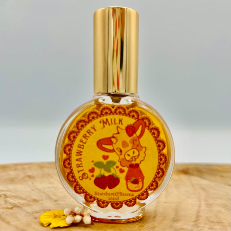 Puede incluir: Una peque&ntilde;a botella de vidrio con una tapa dorada y una etiqueta amarilla. La etiqueta presenta una vaca de dibujos animados con una fresa en la cabeza y el texto "Strawberry Milk". La botella es de 15 ml y est&aacute; etiquetada como "Stardust & Stems".