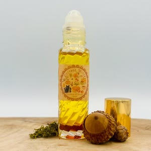 May include: A clear glass roll-on bottle filled with golden liquid, featuring a label with the text Cottage Witch Stardust & Stems 10ml. The bottle has a white plastic roller ball top, and is accompanied by a gold cap and a brown acorn cap.