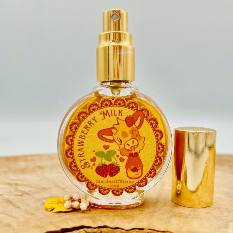 Puede incluir: Una peque&ntilde;a botella de vidrio con una tapa dorada y una etiqueta que dice "Strawberry Milk". La etiqueta presenta una vaca de dibujos animados con una fresa en la cabeza. La botella es de 15 ml y est&aacute; hecha por Stardust & Stems.