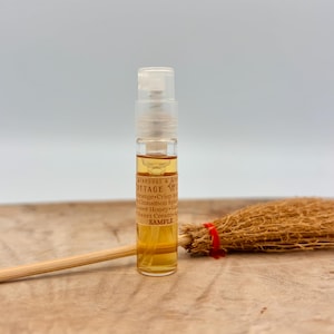 May include: A small clear glass spray vial filled with amber liquid, featuring a label that reads STARDUST & STONES COTTAGE WICK, Orange, Crisp Apple, Cinnamon Spice, Flower Honey, Vanilla, Sweet Cream, SAMPLE. A miniature straw broom lies horizontally.
