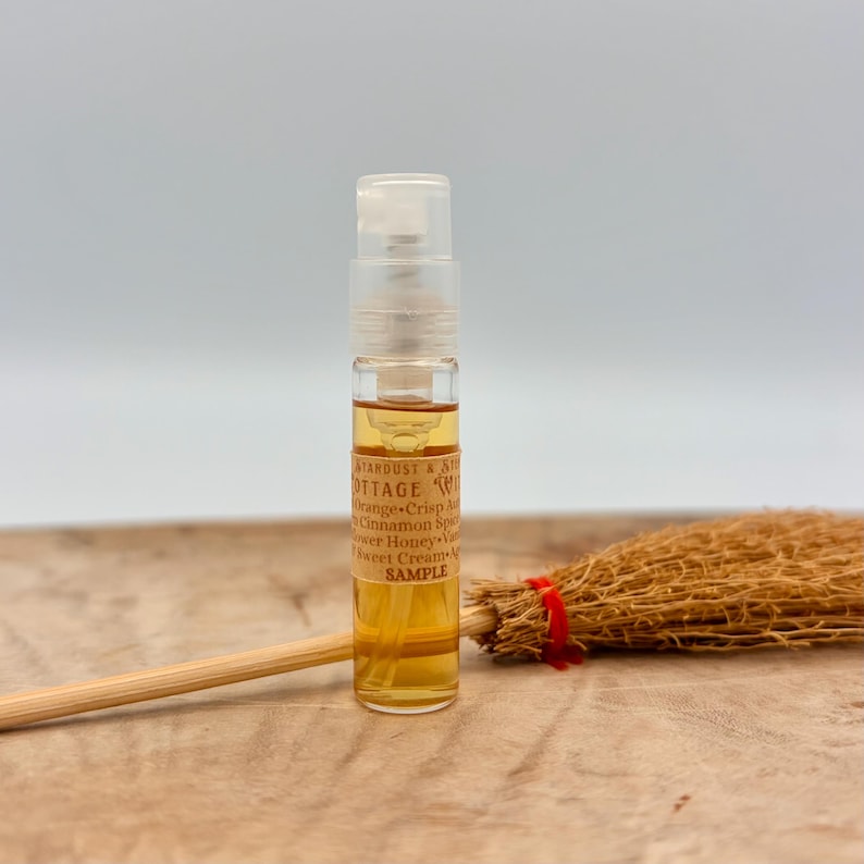 May include: A small clear glass spray vial filled with amber liquid, featuring a label that reads STARDUST & STONES COTTAGE WICK, Orange, Crisp Apple, Cinnamon Spice, Flower Honey, Vanilla, Sweet Cream, SAMPLE. A miniature straw broom lies horizontally.