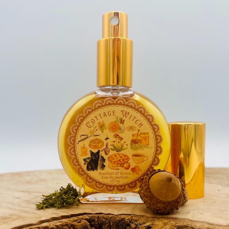 May include: A round glass perfume bottle with a gold spray nozzle and cap, containing a golden liquid. The label features the text Cottage Witch and illustrations of a black cat, mushrooms, a pie, and a candle, with an acorn and moss nearby.