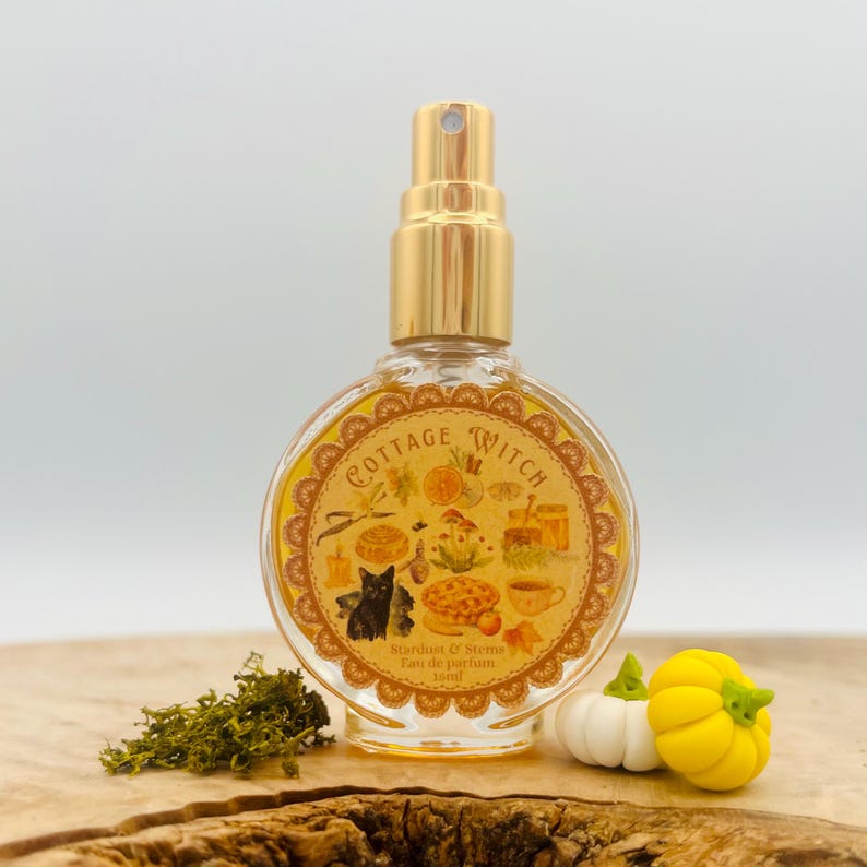 May include: A round glass perfume bottle with a gold spray nozzle, containing a yellow liquid. The label reads Cottage Witch, Stardust & Stems, Eau de parfum, 16ml, and features illustrations of a black cat, mushrooms, and a pie.