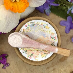 May include: A small, round, brown container with a white label that says "Stardust & Stems Petal Powder Oh, Apothecary Shimmer 0.5 oz". The container has a pink and white spoon with a wooden handle.